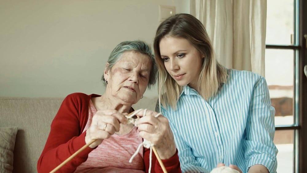 Una cuidadora con una señora mayor de edad tejiendo 