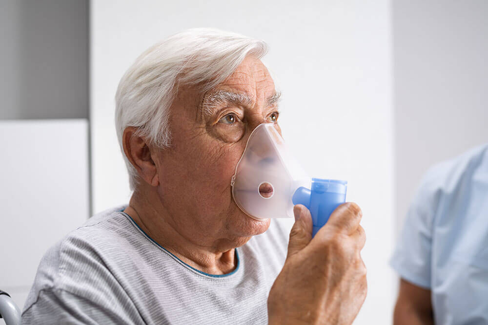 Anciano inhala oxígeno con un oxigenador que lleva en la mano.