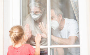 Abuelos con mascarilla saludando a su nieta tras la ventana