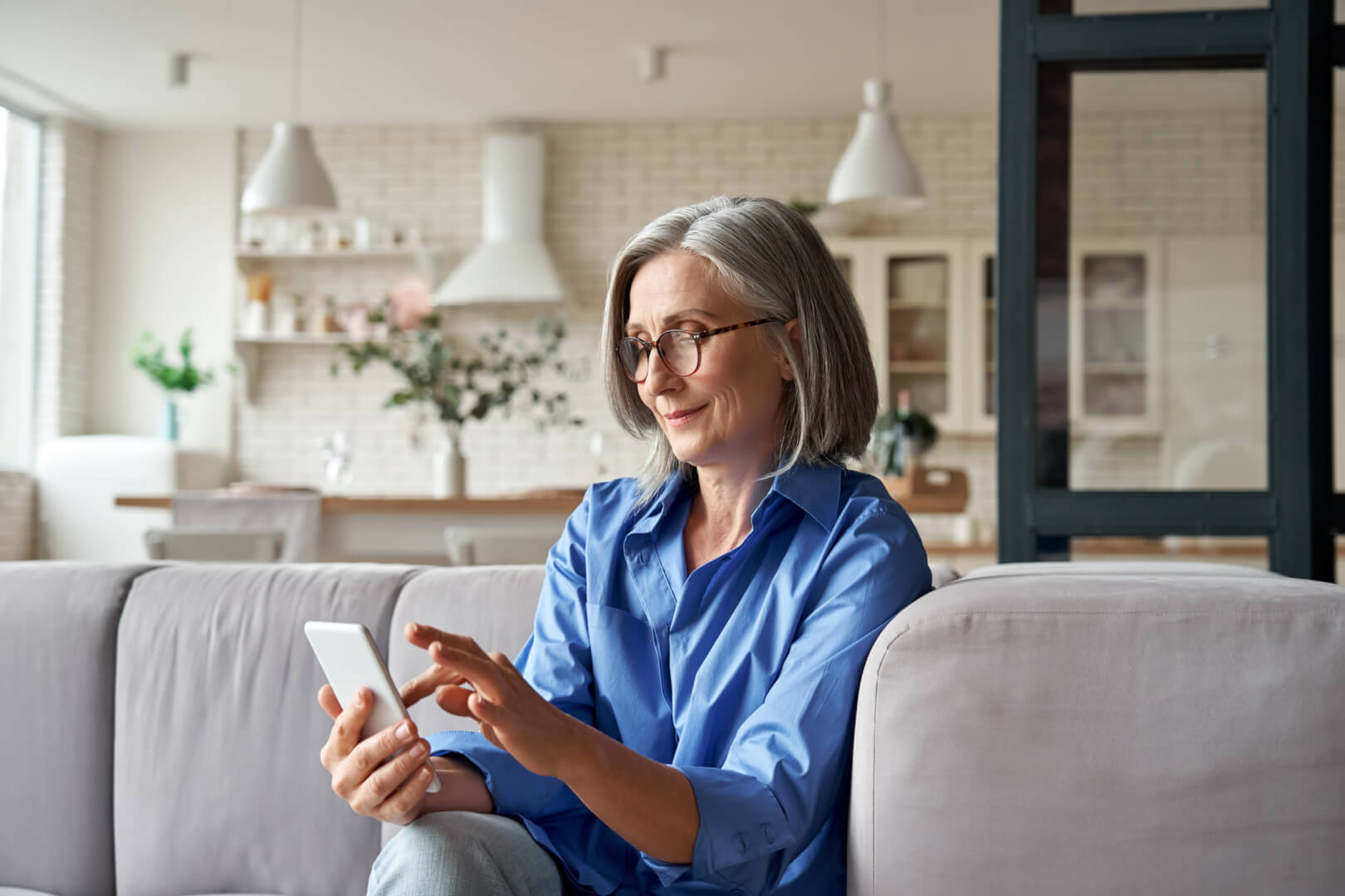 Una mujer de la tercera edad sentada en el sofá de su casa, con gafas mira el móvil.