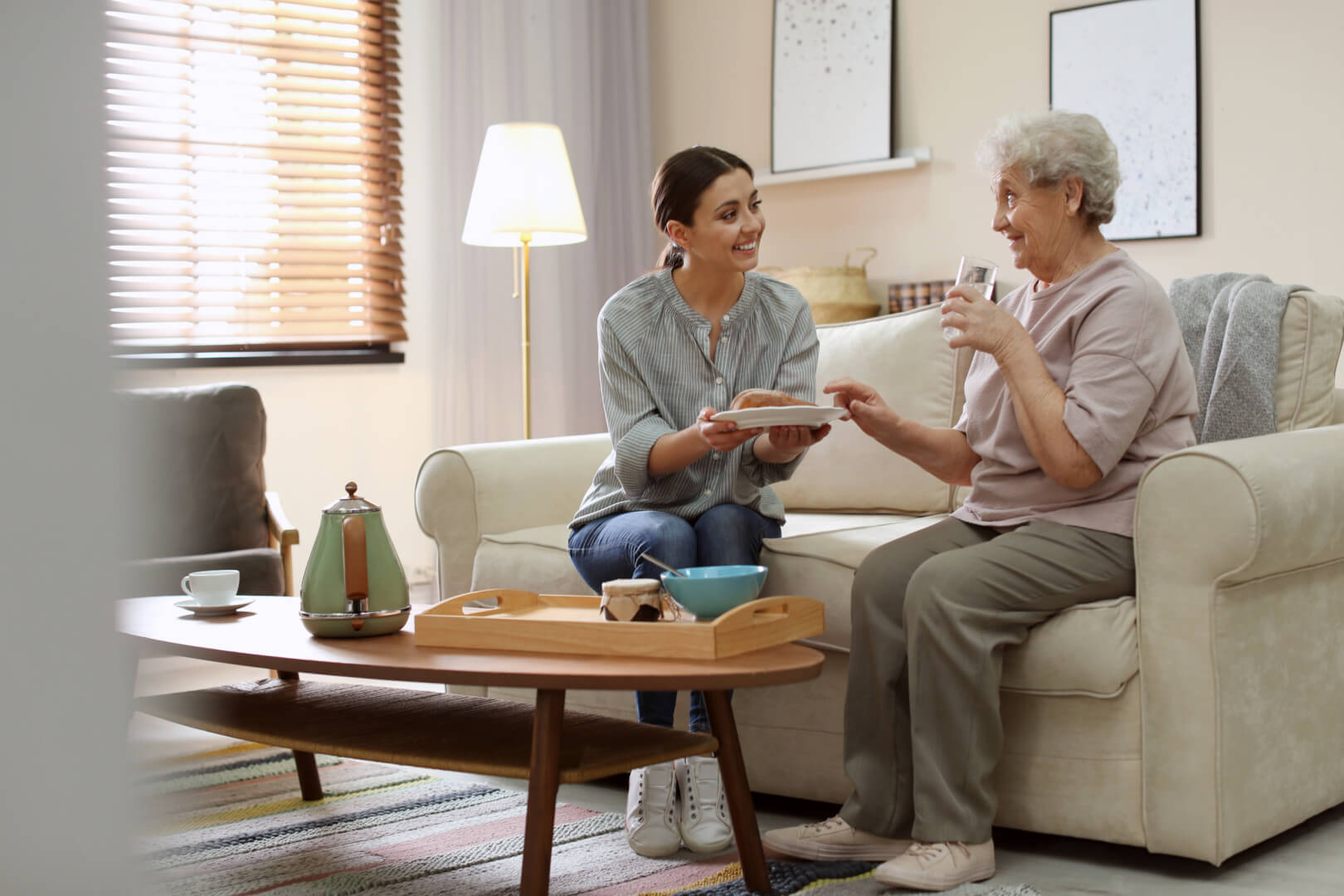 En la ilustración están dos personas, una mujer joven a la izquierda y una señora de edad avanzada, sentadas en un sofá, mientras hablan y la chica joven le ofrece a la persona mayor un plato de comida, en un desayuno