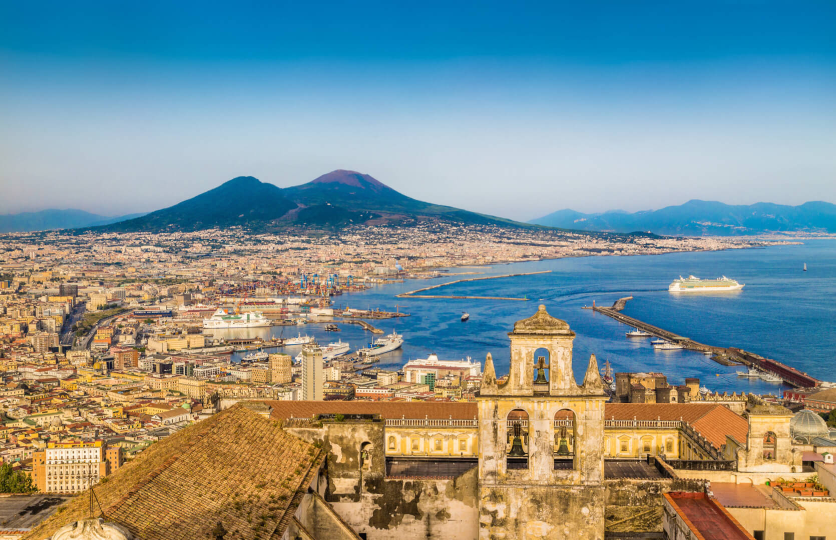 Aparece una vista panorámica de la ciudad de Nápoles en Italia con el mar a la derecha y las montañas al fondo.