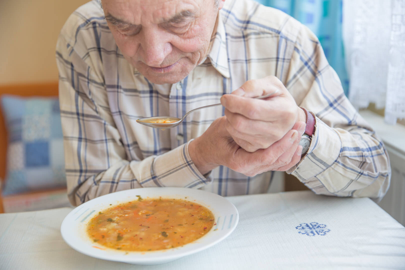 En la imagen aparece un señor mayor que tiene que sujetarse la mano con la otra mano mientras come un plato de sopa por el temblor que le produce el párkinson. 