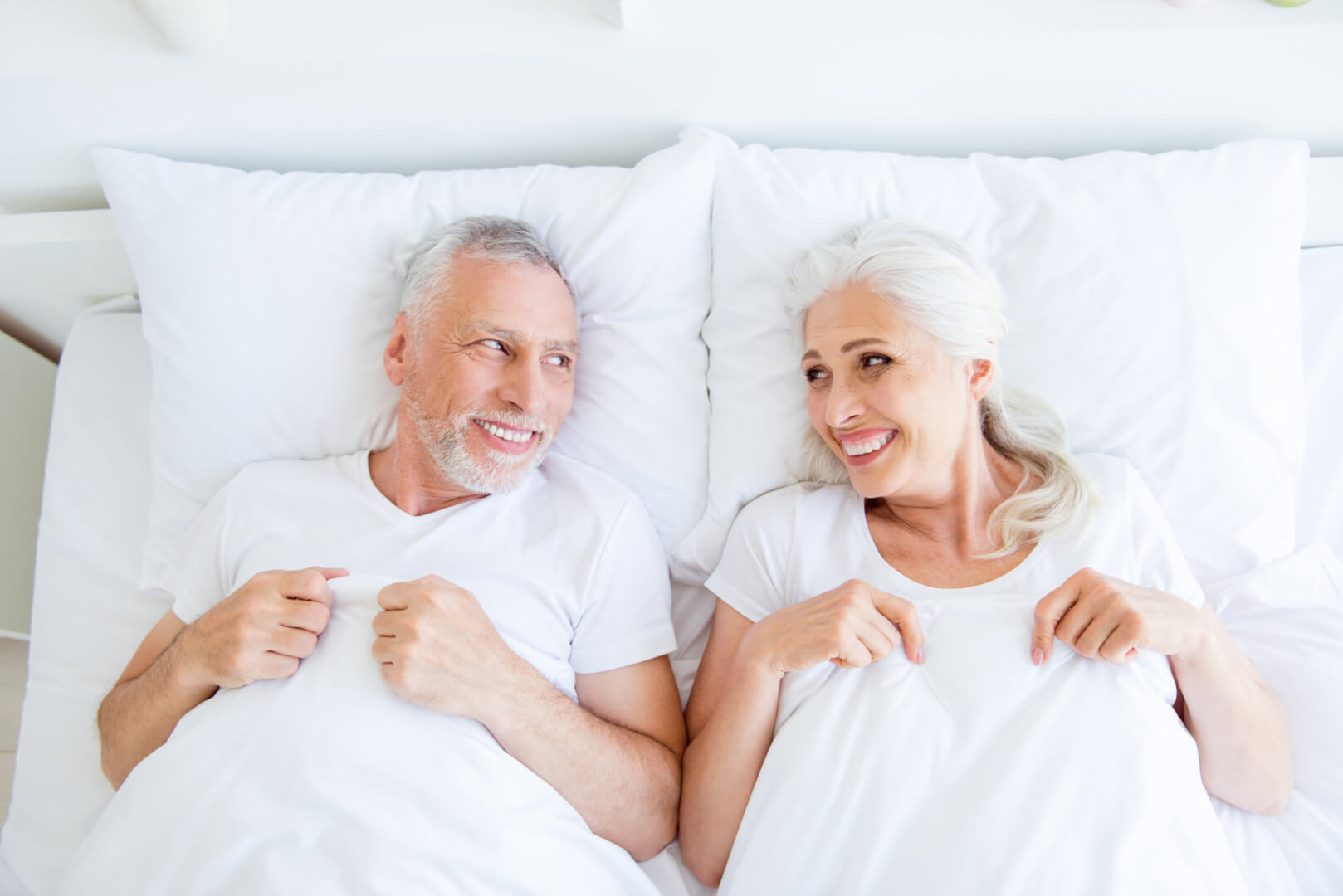 En la imagen se muestra a dos personas mayores, un matrimonio de un varón y una mujer en la cama mirándose y sonriéndose. Hacer el amor a partir de los 60 tiene múltiples beneficios emocionales. 