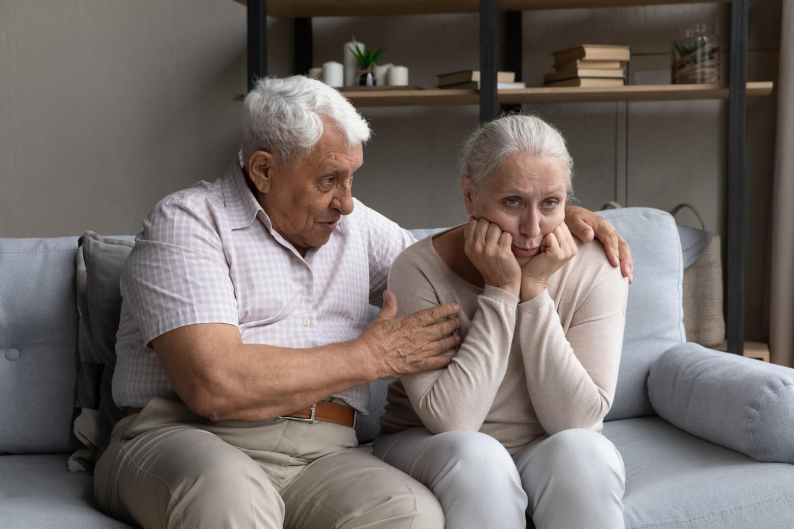 Pareja de personas mayores que sufren Parkinson sentados en el sofá de su casa mientras el hombre mayor consuela y abraza a su mujer.