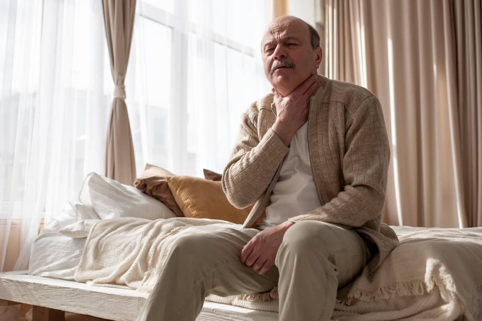 Un señor mayor está sentado en su cama mientras se queja de una dolencia de tiroides en el cuello. 