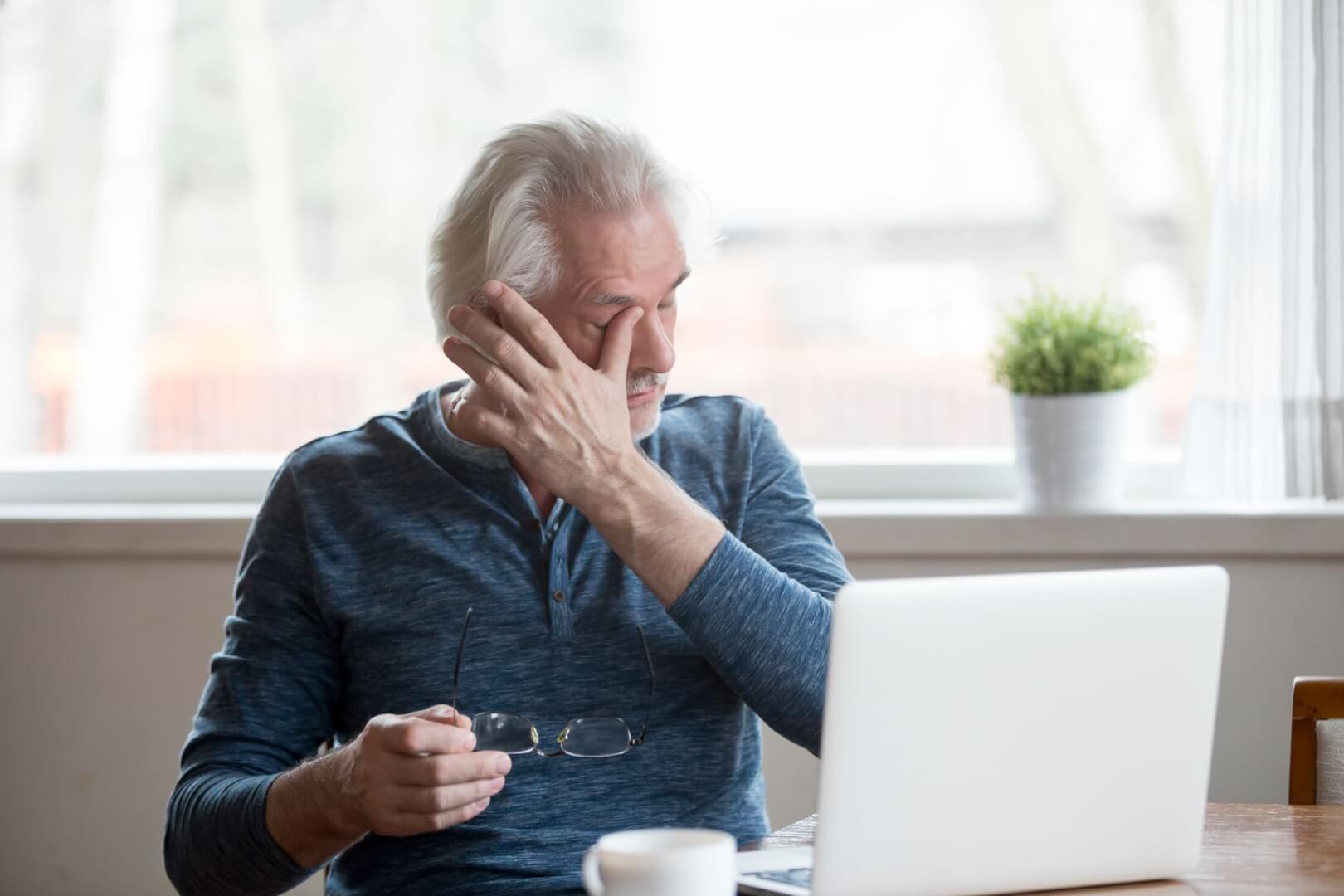 Un señor mayor se frota el ojo ante una molestia mientras escribe en el ordenador. 